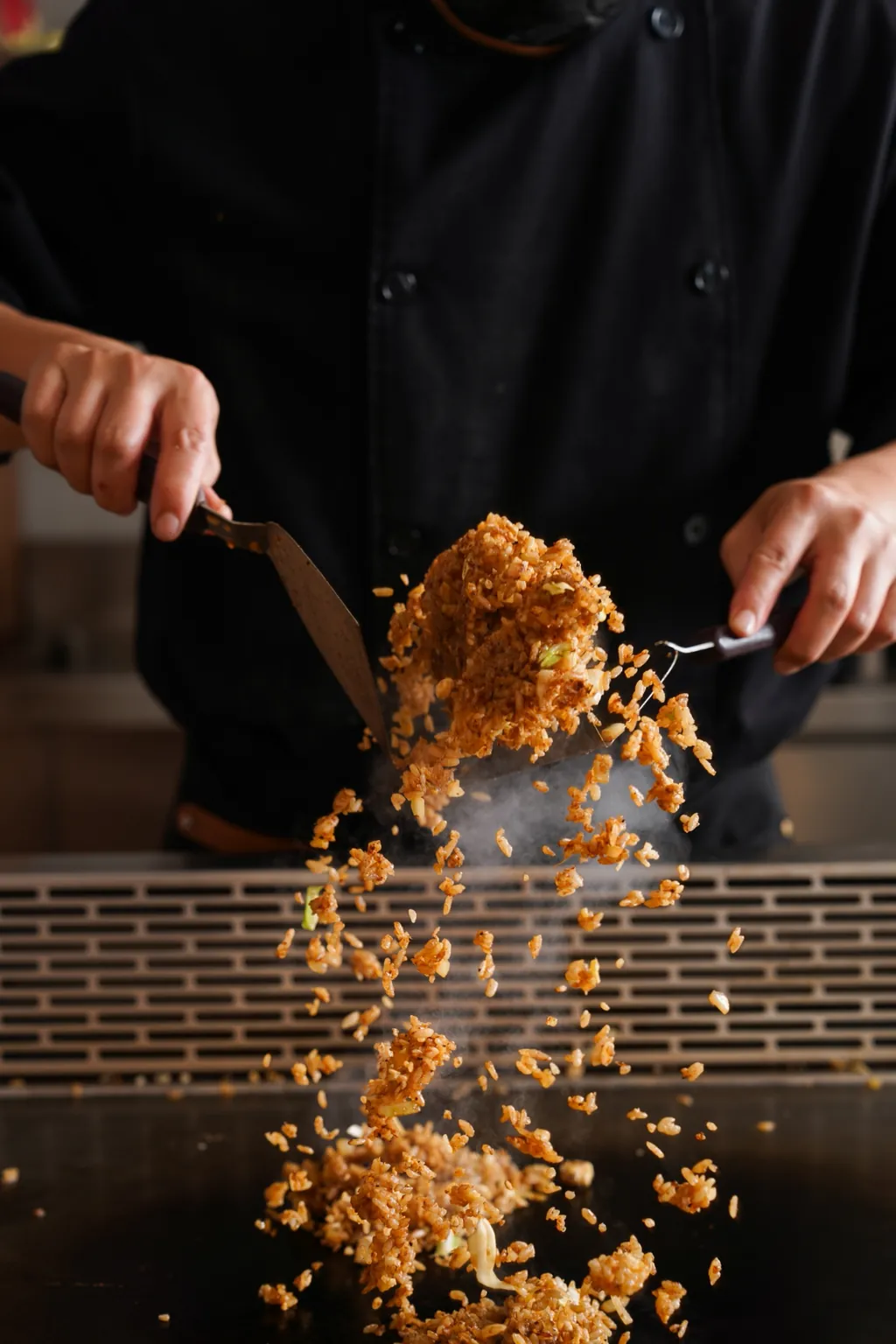 A chef tossing fried rice on the griddle with two spatulas, grains flying through the air